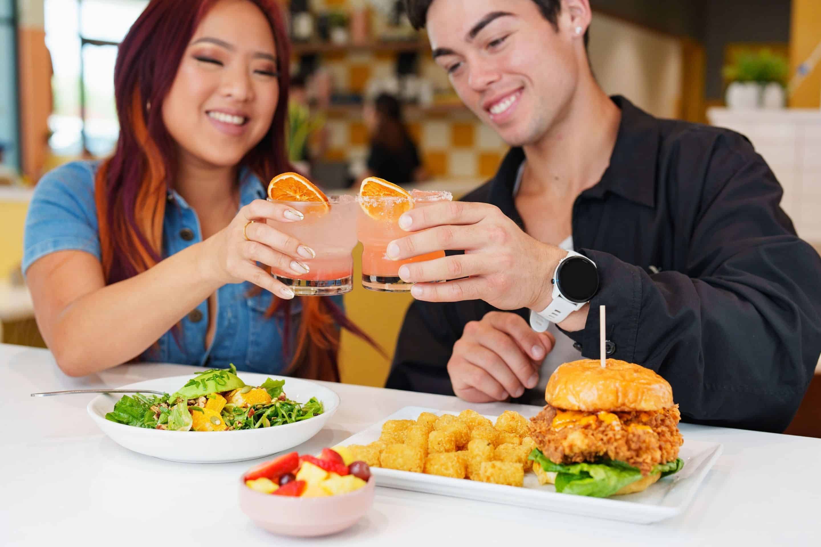 Couple raising their cocktails during brunch at Over Easy, chicken sandwich and salad entrees are on the table